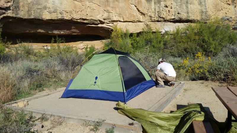 A man setting up a green tent next to rock wall at a small Chaco Canyon camping site.