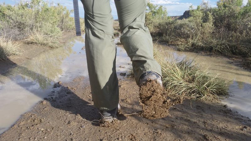 A woman wearing green pants lifting her right foot to reveal caked mud on the outer sole of her hiking boot.