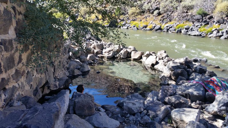 A clear pool of water with a man soaking in it on the edge of a river near Taos, New Mexico.