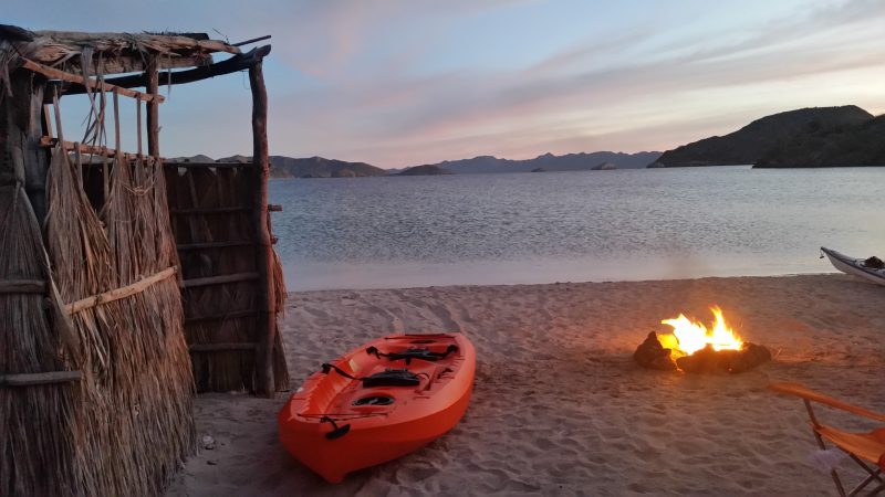 An orange kayak on the beach by a campfire with purple sunset in the background.