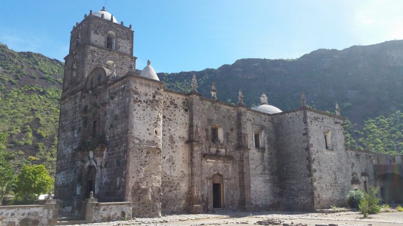 A large hand cut stone mission church with white domes surrounded by mountains.