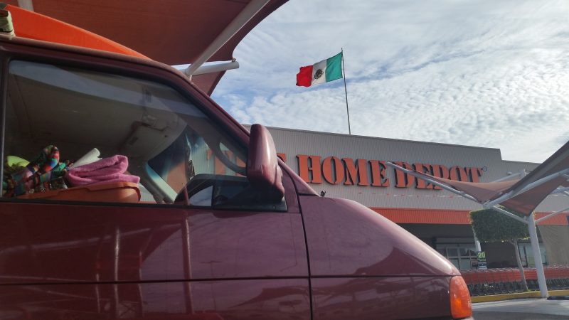 A maroon Volkswagen van outside of Home Depot in La Paz, Baja California which has a Mexican flag blowing in the wind on its roof.