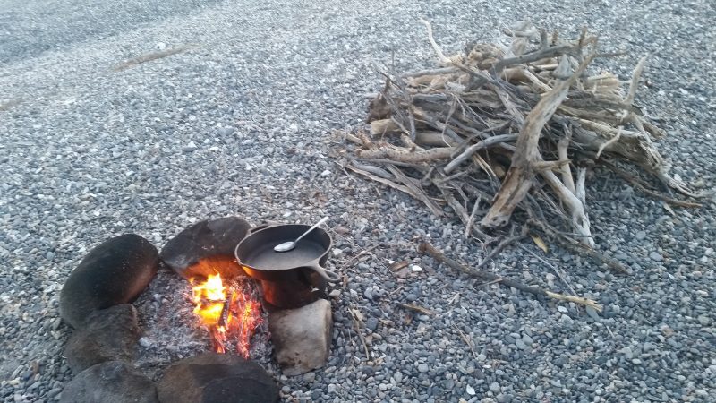 Baja camping on the beach with a fire and a skillet and a pile of fire wood nearby.