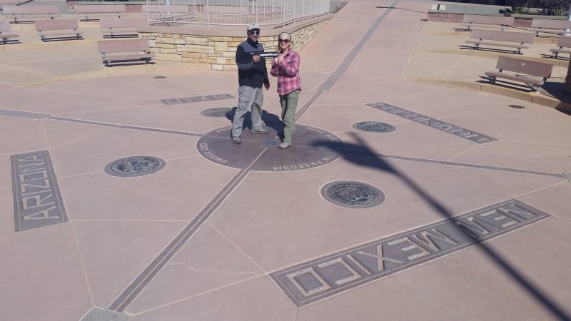 A man and woman holding a bumper sticker at the Four Corners Monument, a spot popular with tourists on road trips in New Mexico.