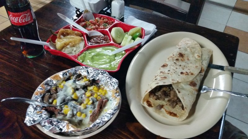 A table with a giant burrito, baked potato, salsa platter and a bottle of Coke from a popular family-style restaurant in Baja California.