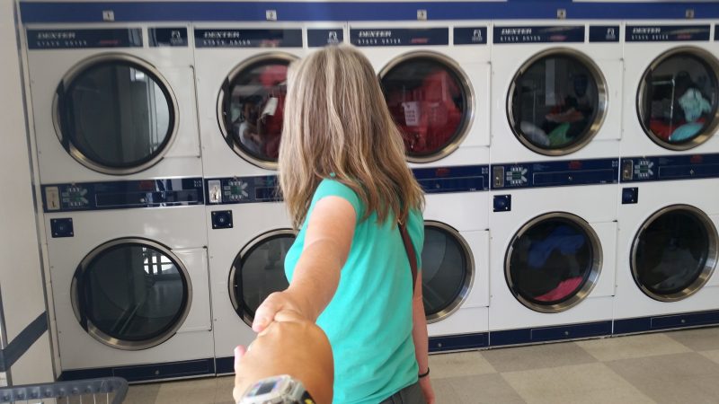 Woman in green T shirt leading man by the hand to washing machines in a laundromat.