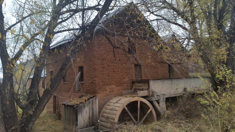 An old adobe mill building with water wheel surrounded by autumn trees.