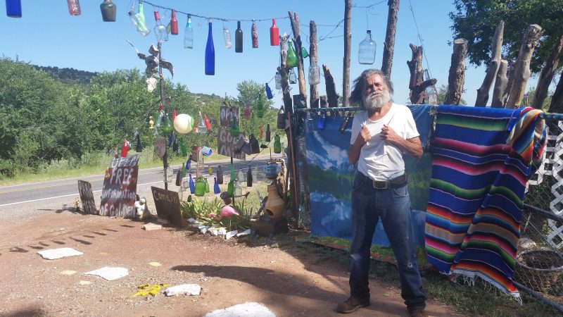 A man with a beard wearing blue jeans and a white shirt standing in front of bottles hanging from strings on New Mexico's famous Turquoise Trail.