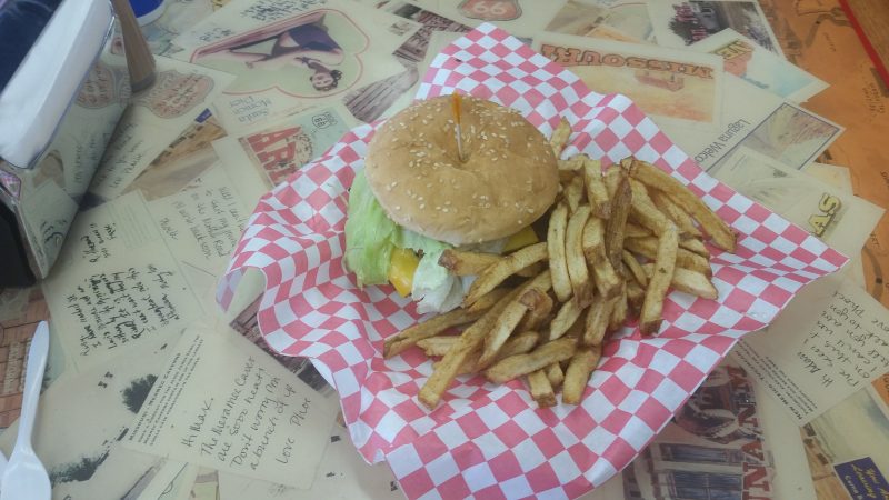 A green chile cheeseburger in a basket with french fries on the side, sitting on a table decorated with vintage postcards at Laguna Burger in Albuquerque.
