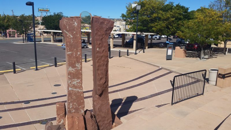 Two large rough-hewn red stone obelisks with a glass circle between them acting as a sun dial.