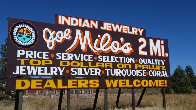 A brown sign with yellow and white lettering advertising Joe Milo's Trading Post, a popular weekend excursion in New Mexico for shoppers in search of native crafts.