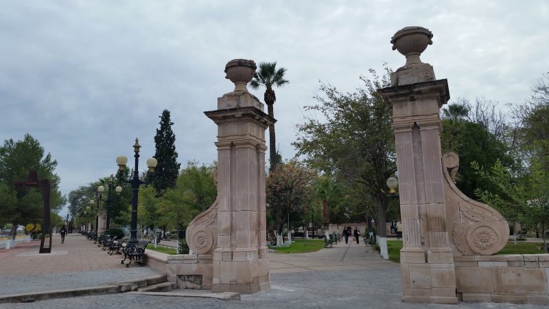 Two stone pillars at the entrance of a park with benches and a tall palm tree.