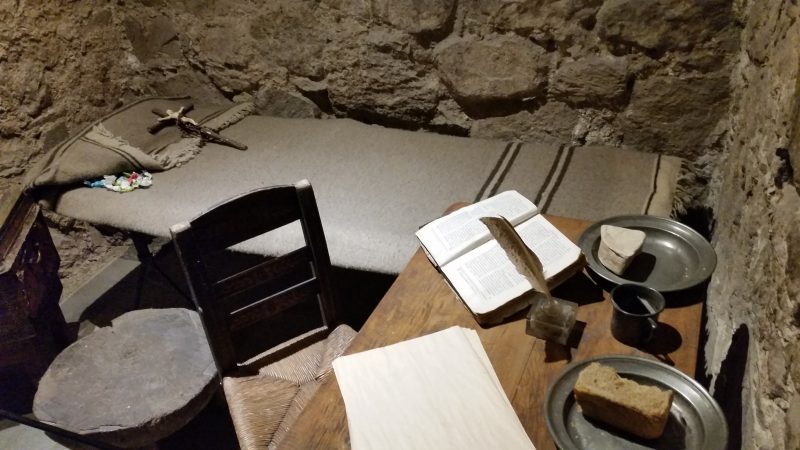 An old wooden chair and desk with a book and a plate of food on it and a bed in the background with a small crucifix on it.