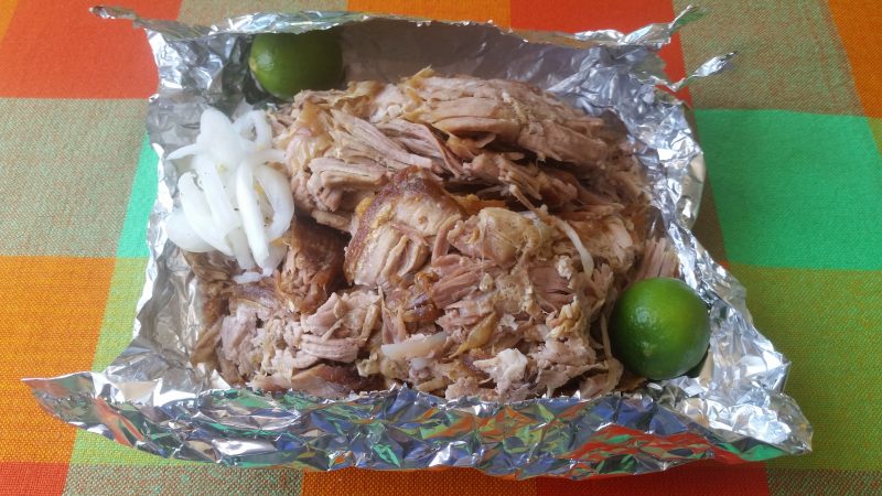 A platter of carnitas, or shredded pork, on a large piece of aluminum foil with limes and onions garnishing it.