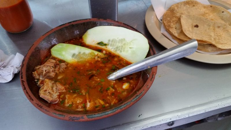 A budget backpacking meal in Mexico of a bowl of beef soup with red broth and a plate of tortillas.