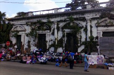 An old building in San Salvador overgrown with vegetation and with several types of goods being sold in front of it.