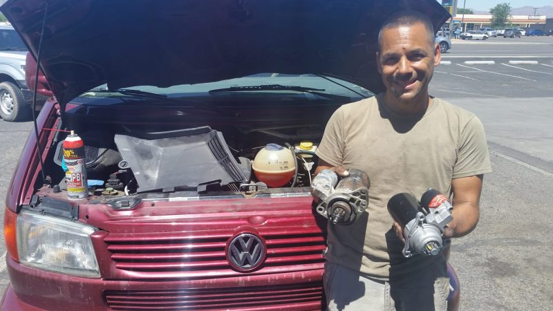 A man holding two starter motors in front of a burgundy Volkswagen van with the hood up.