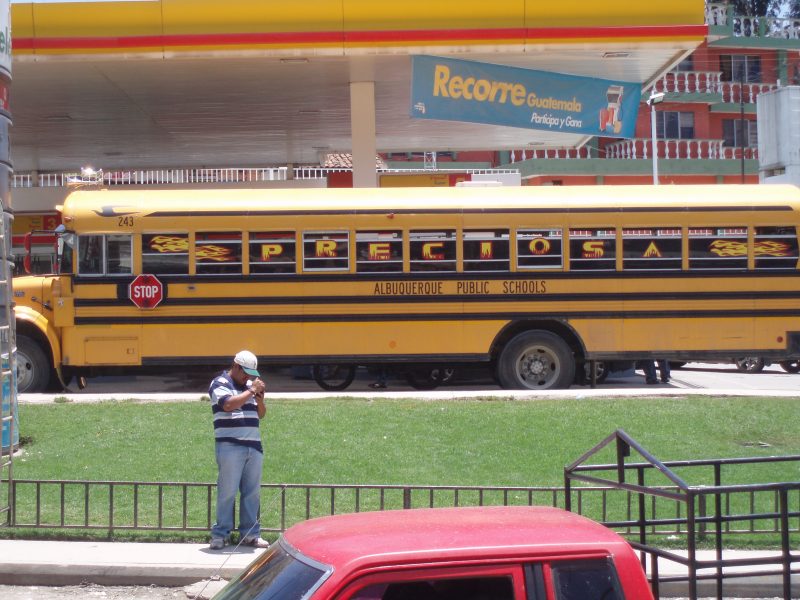 Man in baseball hat lighting a cigarette in front of a yellow school bus.