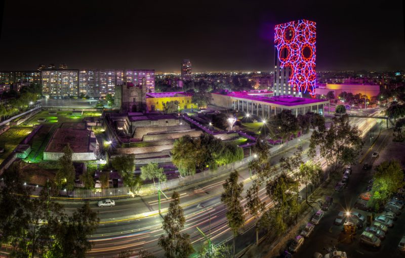 Overexposed view of historic square in Mexico City with a multi-coloured modern building lit up at night.