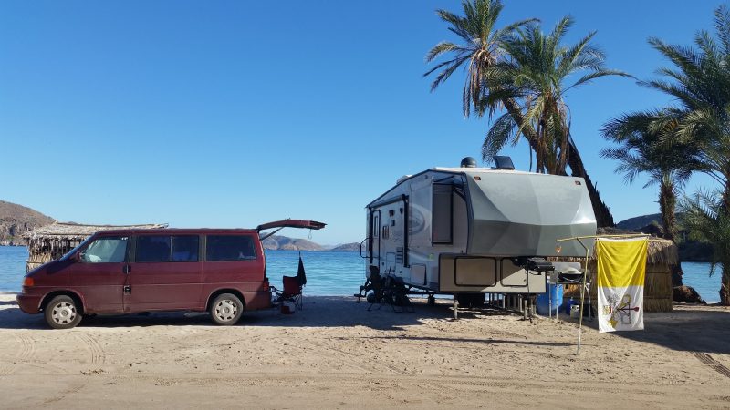 A burgundy Volkswagen van and an RV Baja California camping on a white sand beach with a palm tree in the background.