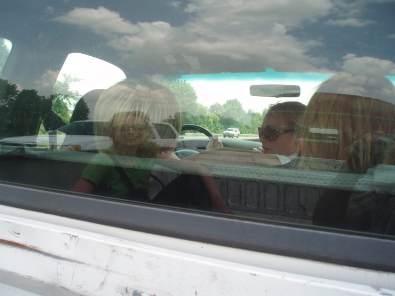 A reflection of a smiling woman in the glass of a windshield.